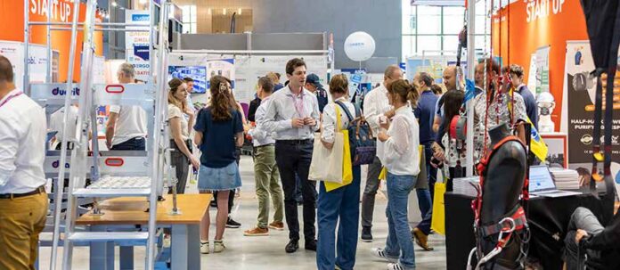 Un groupe de personnes se promène et discute parmi les stands d'un salon de la création d'entreprise. Des bannières et des présentoirs colorés entourent la foule, et certains participants portent des sacs fourre-tout et des badges nominatifs.