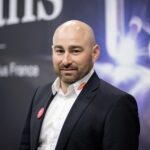 Un homme chauve à la barbe taillée, vêtu d'un costume sombre et d'une chemise blanche, se tient à l'intérieur devant un arrière-plan flou comportant du texte et des formes abstraites.