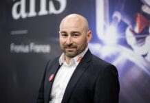 Soudage : la cobotique doit changer de dimension Un homme chauve à la barbe taillée, vêtu d'un costume sombre et d'une chemise blanche, se tient à l'intérieur devant un arrière-plan flou comportant du texte et des formes abstraites.