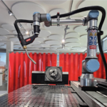 Un bras de soudage robotisé est positionné au-dessus d'une table de travail en métal perforé dans un environnement industriel, avec des étiquettes d'avertissement de sécurité visibles sur le bras et un rideau de protection rouge à l'arrière-plan.