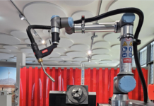 Un bras de soudage robotisé est positionné au-dessus d'une table de travail en métal perforé dans un environnement industriel, avec des étiquettes d'avertissement de sécurité visibles sur le bras et un rideau de protection rouge à l'arrière-plan.