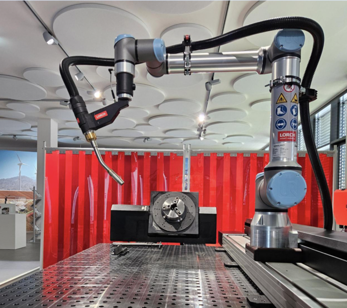 Un bras de soudage robotisé est positionné au-dessus d'une table de travail en métal perforé dans un environnement industriel, avec des étiquettes d'avertissement de sécurité visibles sur le bras et un rideau de protection rouge à l'arrière-plan.