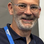 Un homme aux cheveux courts, aux lunettes et à la barbe grise taillée porte un polo bleu marine et un cordon bleu portant le mot "Linde". Il sourit et se tient devant un fond gris.