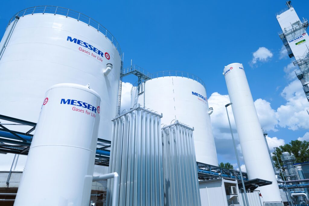 Grands réservoirs et bouteilles de gaz industriels blancs portant le logo "Messer Gases for Life", placés en extérieur sous un ciel bleu avec des nuages. Des tuyaux métalliques et d'autres structures industrielles sont visibles.