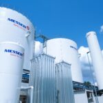 Grands réservoirs et bouteilles de gaz industriels blancs portant le logo "Messer Gases for Life", placés en extérieur sous un ciel bleu avec des nuages. Des tuyaux métalliques et d'autres structures industrielles sont visibles.