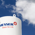 Un réservoir industriel blanc portant le logo "Messer Gases for Life" est représenté sur fond de ciel bleu avec quelques nuages blancs.
