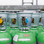 Plusieurs bouteilles de gaz de soudage vertes et argentées sont alignées à l'extérieur. Chaque bouteille est munie d'un robinet au sommet, et certaines sont munies d'étiquettes en plastique jaunes et bleues. Les étiquettes et les codes-barres sont clairement visibles sur les bouteilles.