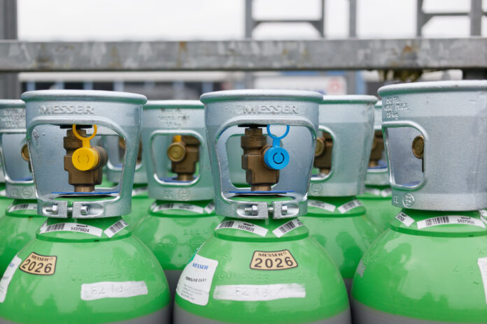 Plusieurs bouteilles de gaz de soudage vertes et argentées sont alignées à l'extérieur. Chaque bouteille est munie d'un robinet au sommet, et certaines sont munies d'étiquettes en plastique jaunes et bleues. Les étiquettes et les codes-barres sont clairement visibles sur les bouteilles.