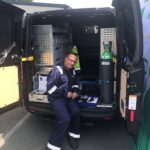 Une personne portant un uniforme bleu foncé avec des bandes réfléchissantes est assise sur le bord d'une camionnette ouverte. La camionnette est équipée d'étagères, de matériel et d'une grande bouteille de gaz vert. L'environnement semble être un parking.