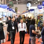Les visiteurs déambulent et discutent dans un hall d'exposition animé, bordé de part et d'autre par les stands des entreprises. Des panneaux au-dessus des stands indiquent les noms des entreprises et les numéros des halls. L'ambiance est professionnelle et axée sur les affaires.