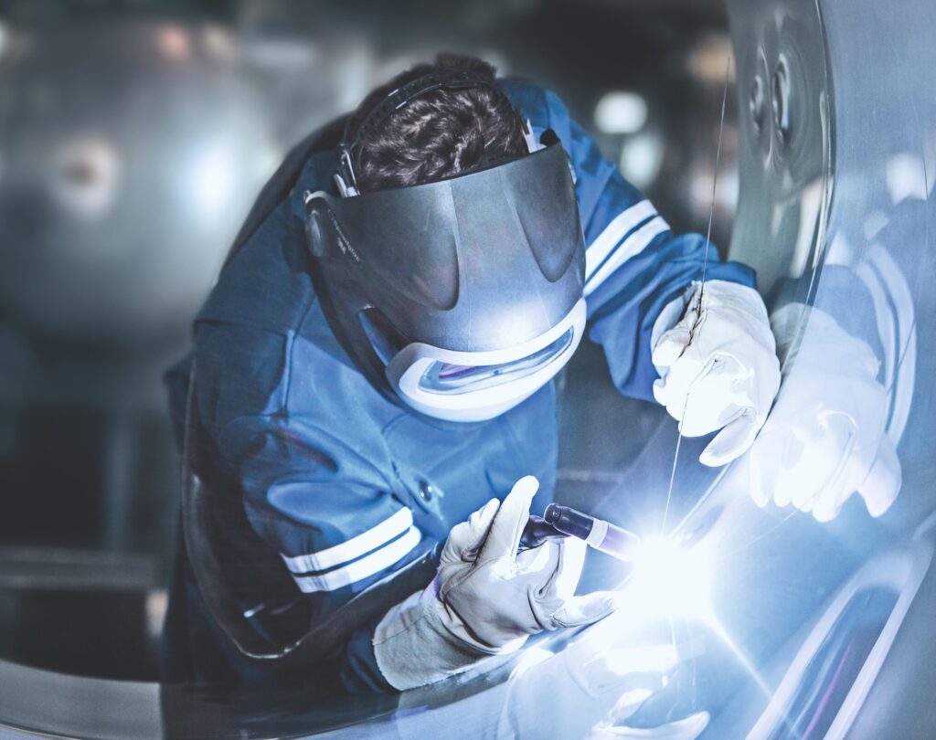 Une personne portant des vêtements de protection, des gants et un écran facial utilise une torche de soudage pour travailler sur une surface métallique dans un environnement industriel.