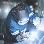 Une personne portant des vêtements de protection, des gants et un écran facial utilise une torche de soudage pour travailler sur une surface métallique dans un environnement industriel.