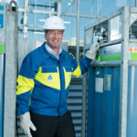 Un homme portant un casque blanc, des gants et une veste de sécurité bleue et jaune se tient entre deux grands cylindres de gaz industriels à l'extérieur d'un bâtiment et sourit à la caméra.