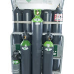 Un ensemble de six grandes bouteilles de gaz comprimé et de trois plus petites portant des étiquettes vertes et noires, fixées verticalement dans un rack de stockage métallique. Les étiquettes de sécurité et d'identification sont visibles sur le casier et les bouteilles.