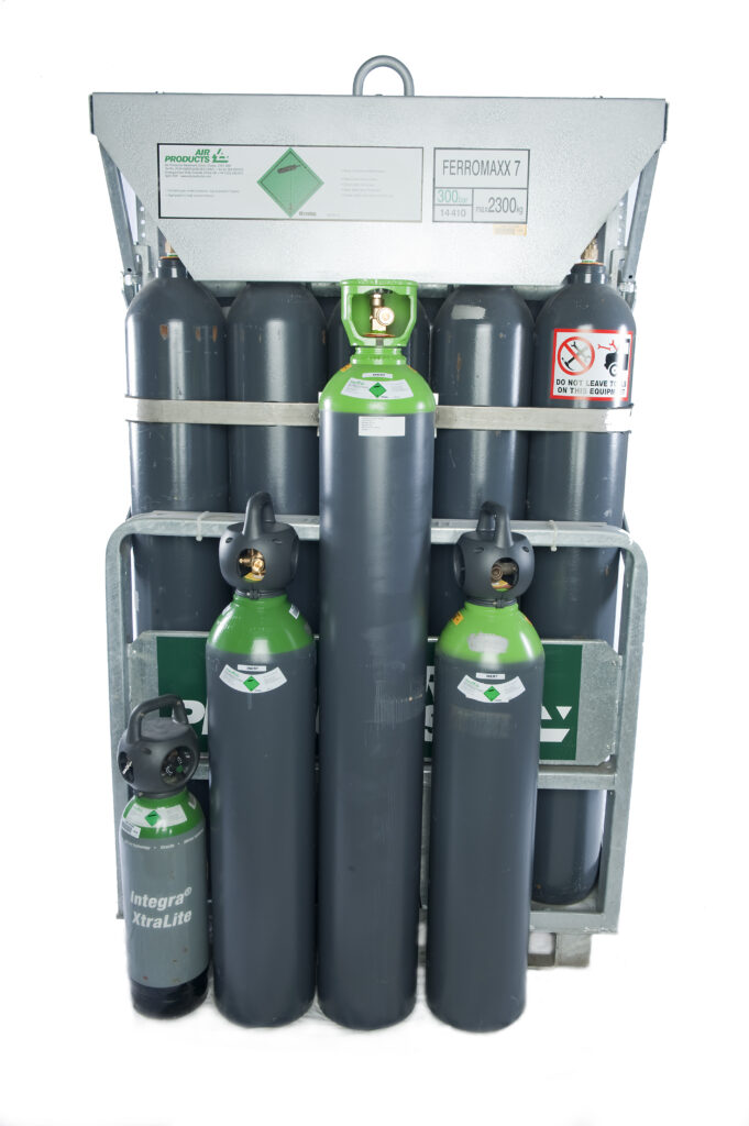 Un ensemble de six grandes bouteilles de gaz comprimé et de trois plus petites portant des étiquettes vertes et noires, fixées verticalement dans un rack de stockage métallique. Les étiquettes de sécurité et d'identification sont visibles sur le casier et les bouteilles.