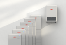 Fronius présente la batterie haute tension Reserva Pro et son intégration GEN24 Plus / Verto Plus Six unités de stockage d'énergie blanches sont alignées sur le sol, tandis qu'un appareil mural est placé à droite. Tous les appareils arborent le même logo de marque rouge et blanc ainsi que des voyants lumineux bleus.