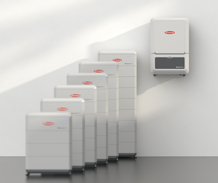 Six unités de stockage d'énergie blanches sont alignées sur le sol, tandis qu'un appareil mural est placé à droite. Tous les appareils arborent le même logo de marque rouge et blanc ainsi que des voyants lumineux bleus.