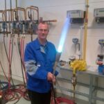 Un homme vêtu d'une veste de travail bleue tient un chalumeau allumé avec une flamme bleue dans un environnement industriel. Des tuyaux, des vannes, des compteurs et des équipements sont visibles sur les murs et l'établi derrière lui.