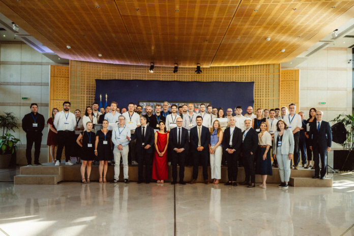Un grand groupe de personnes en tenue professionnelle et habillée pose pour une photo de groupe dans un espace intérieur moderne et bien éclairé, avec un fond bleu derrière elles. Certaines personnes portent des badges nominatifs.