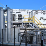 Un site industriel composé de tuyaux métalliques, d'un périmètre clôturé et d'un bâtiment blanc portant l'inscription « Lhyfe ». On y voit des rampes jaunes le long d'un escalier ainsi que divers équipements industriels, le tout sous un ciel bleu limpide.
