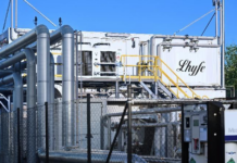 Un site industriel composé de tuyaux métalliques, d'un périmètre clôturé et d'un bâtiment blanc portant l'inscription « Lhyfe ». On y voit des rampes jaunes le long d'un escalier ainsi que divers équipements industriels, le tout sous un ciel bleu limpide.
