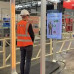 Une personne portant un équipement de protection individuelle (EPI) — un casque de chantier blanc et un gilet de sécurité orange — se tient debout dans un portique de détection JO-Safe, face à un écran qui affiche son image ainsi que le message « Accès refusé » accompagné d’une croix rouge, indiquant que l’accès lui est refusé.