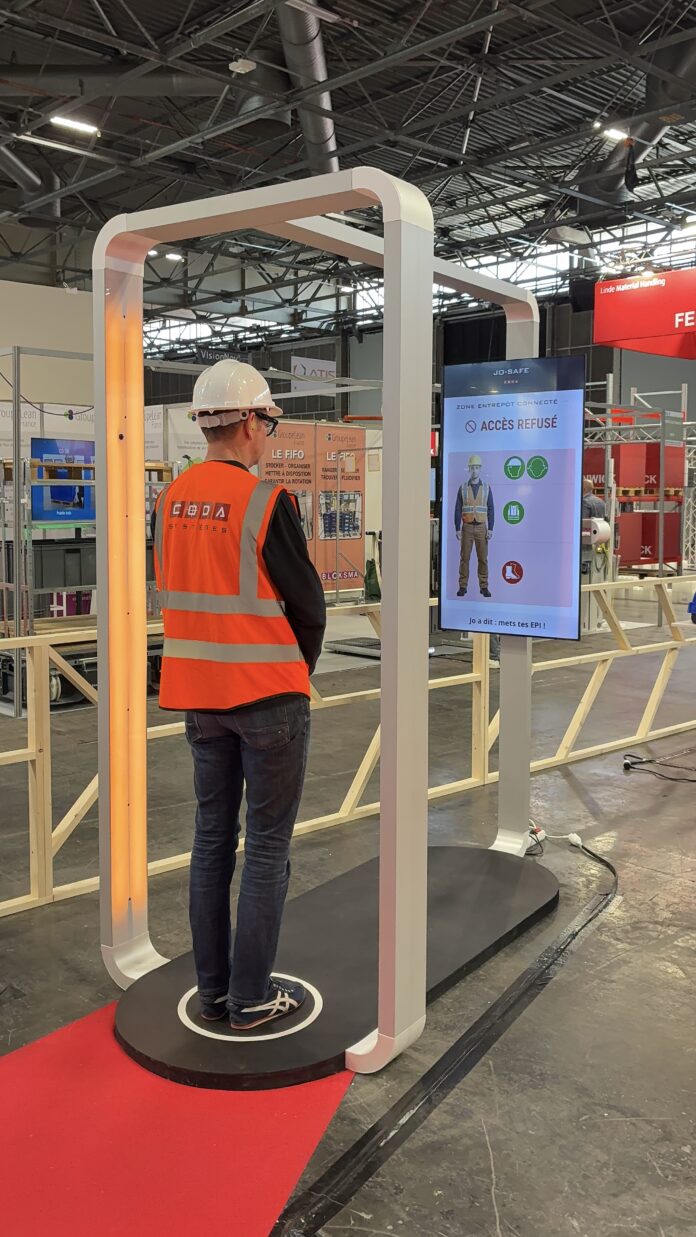 Une personne portant un équipement de protection individuelle (EPI) — un casque de chantier blanc et un gilet de sécurité orange — se tient debout dans un portique de détection JO-Safe, face à un écran qui affiche son image ainsi que le message « Accès refusé » accompagné d’une croix rouge, indiquant que l’accès lui est refusé.