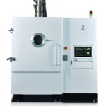 Une grande machine industrielle blanche conçue pour l'impression 3D métal, dotée d'une fenêtre d'observation circulaire, d'un panneau de commande avec écran et boutons, ainsi que de voyants lumineux sur le dessus. La machine est posée sur une surface réfléchissante, devant un fond blanc.