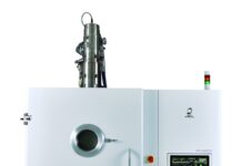 Une grande machine industrielle blanche conçue pour l'impression 3D métal, dotée d'une fenêtre d'observation circulaire, d'un panneau de commande avec écran et boutons, ainsi que de voyants lumineux sur le dessus. La machine est posée sur une surface réfléchissante, devant un fond blanc.