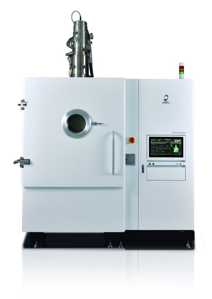 Une grande machine industrielle blanche conçue pour l'impression 3D métal, dotée d'une fenêtre d'observation circulaire, d'un panneau de commande avec écran et boutons, ainsi que de voyants lumineux sur le dessus. La machine est posée sur une surface réfléchissante, devant un fond blanc.