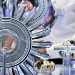 Gros plan sur une roue de turbine métallique brillante, dotée de plusieurs aubes incurvées, exposée et entourée d'autres pièces mécaniques et d'équipements techniques, sur fond industriel flou.
