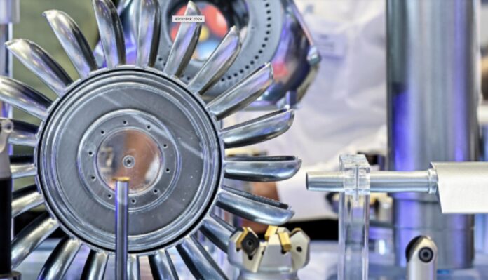 Gros plan sur une roue de turbine métallique brillante, dotée de plusieurs aubes incurvées, exposée et entourée d'autres pièces mécaniques et d'équipements techniques, sur fond industriel flou.