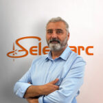 Un homme aux bras croisés, portant une barbe grise, se tient debout devant un mur orné du logo orange « Sele » et « arc », vêtu d'une chemise bleu clair.