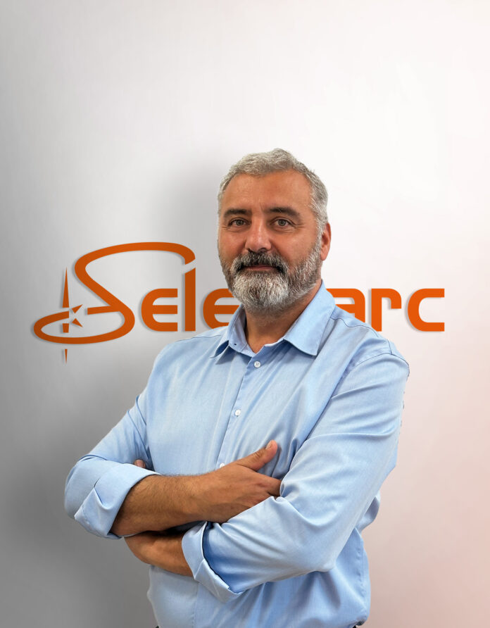 Un homme aux bras croisés, portant une barbe grise, se tient debout devant un mur orné du logo orange « Sele » et « arc », vêtu d'une chemise bleu clair.