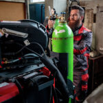 Dans un atelier, un homme règle la vanne d'une grande bouteille de gaz verte, à côté de machines rouges. Il porte une combinaison de travail rouge et noire ainsi que des gants de sécurité. Le décor comprend des équipements industriels et des armoires métalliques.