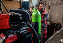 Dans un atelier, un homme règle la vanne d'une grande bouteille de gaz verte, à côté de machines rouges. Il porte une combinaison de travail rouge et noire ainsi que des gants de sécurité. Le décor comprend des équipements industriels et des armoires métalliques.