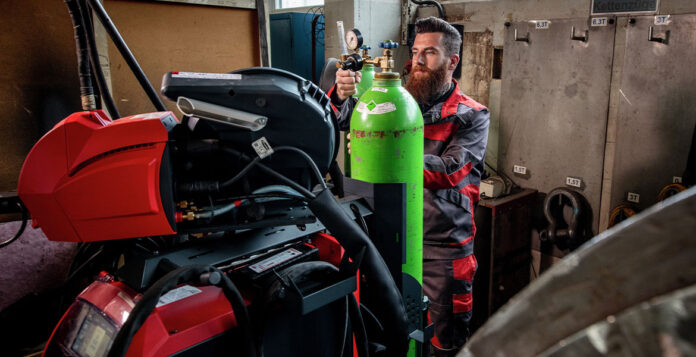 Dans un atelier, un homme règle la vanne d'une grande bouteille de gaz verte, à côté de machines rouges. Il porte une combinaison de travail rouge et noire ainsi que des gants de sécurité. Le décor comprend des équipements industriels et des armoires métalliques.