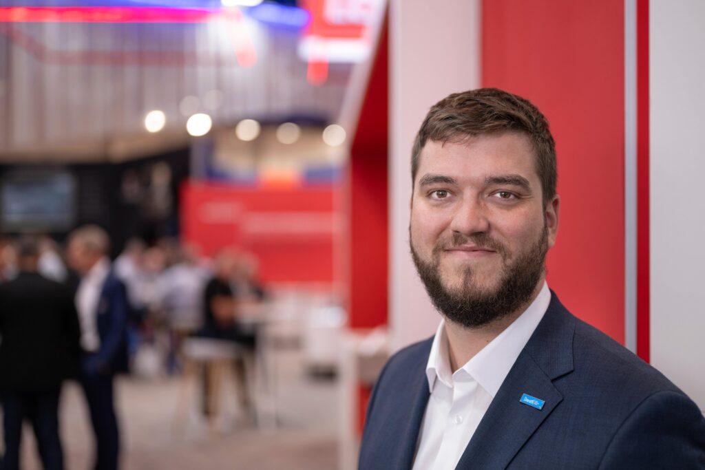 Un homme barbu, vêtu d’un costume et d’une chemise blanche, se tient à l’intérieur et sourit à la caméra. L’arrière-plan est flou et laisse entrevoir des personnes en tenue professionnelle ainsi qu’un décor rouge, ce qui suggère un événement professionnel ou une conférence consacrée à l’avenir du soudage et à l’intégration des cobots dans les pratiques modernes de soudage.
