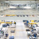 Une vaste halle d'usine bien éclairée, où s'alignent des machines industrielles, des postes de travail et des équipements. Des passerelles et des garde-corps surplombent l'espace de travail. On aperçoit peu d'ouvriers dans la scène.