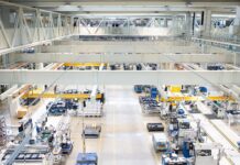 Une vaste halle d'usine bien éclairée, où s'alignent des machines industrielles, des postes de travail et des équipements. Des passerelles et des garde-corps surplombent l'espace de travail. On aperçoit peu d'ouvriers dans la scène.