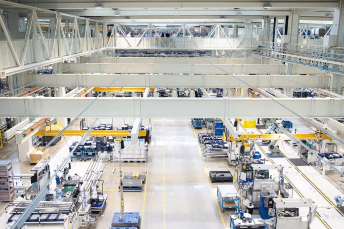 Une vaste halle d'usine bien éclairée, où s'alignent des machines industrielles, des postes de travail et des équipements. Des passerelles et des garde-corps surplombent l'espace de travail. On aperçoit peu d'ouvriers dans la scène.