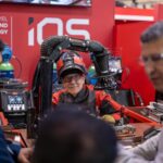 Un homme vêtu d’une tenue de soudeur est assis devant un poste de travail équipé de robots et de cobots, en train de discuter avec trois autres hommes en costume, sur le stand d’un salon technologique où figurent les mentions « NEXT LEVEL HIGH-END TECHNOLOGY » et « iQS ».