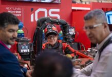 Lorch Schweißtechnik GmbH : soudage manuel, cobots, IA et avenir du soudage en Europe Un homme vêtu d’une tenue de soudeur est assis devant un poste de travail équipé de robots et de cobots, en train de discuter avec trois autres hommes en costume, sur le stand d’un salon technologique où figurent les mentions « NEXT LEVEL HIGH-END TECHNOLOGY » et « iQS ».