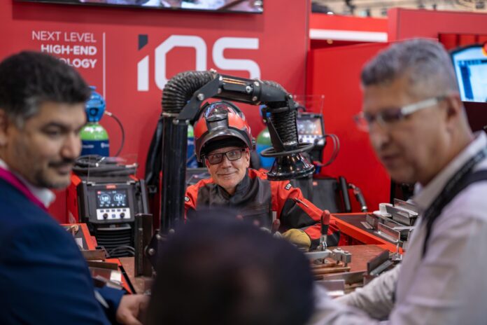 Un homme vêtu d’une tenue de soudeur est assis devant un poste de travail équipé de robots et de cobots, en train de discuter avec trois autres hommes en costume, sur le stand d’un salon technologique où figurent les mentions « NEXT LEVEL HIGH-END TECHNOLOGY » et « iQS ».