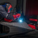 Une personne portant un équipement de sécurité, notamment un casque de soudage et des gants, soude une pièce métallique dans un environnement industriel. On aperçoit à l'arrière-plan une machine à souder portant la marque « Lorch ». Des étincelles bleues jaillissent au point de soudure, soulignant l'importance durable du soudage manuel, alors même que les cobots façonnent l'avenir du soudage.