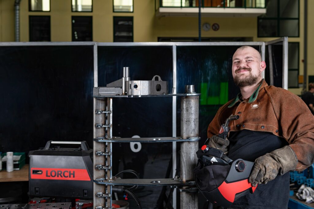 Une personne vêtue d'une tenue de protection se tient debout dans un atelier, souriante, un masque de soudage à la main. On aperçoit du matériel métallique et une machine Lorch, soulignant l'importance du soudage manuel aux côtés d'outils industriels de pointe, avec une cloison sombre en arrière-plan.