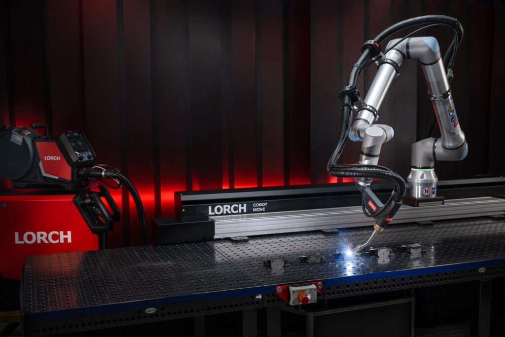 Un bras robotisé équipé d'un dispositif de soudage est à l'œuvre sur un établi métallique, illustrant ainsi la collaboration harmonieuse entre les cobots et les machines industrielles de pointe. L'installation comprend une machine portant l'inscription « Lorch » ainsi que d'autres équipements, le tout dans un atelier à la lumière tamisée, ce qui met en évidence l'intégration de l'automatisation aux procédés traditionnels de soudage manuel.