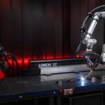 Un bras robotisé équipé d'un dispositif de soudage est à l'œuvre sur un établi métallique, illustrant ainsi la collaboration harmonieuse entre les cobots et les machines industrielles de pointe. L'installation comprend une machine portant l'inscription « Lorch » ainsi que d'autres équipements, le tout dans un atelier à la lumière tamisée, ce qui met en évidence l'intégration de l'automatisation aux procédés traditionnels de soudage manuel.