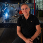 Un homme portant des lunettes et une chemise noire est assis sur un tabouret dans une usine. Derrière lui, un bras robotisé effectue des travaux de soudure sur une table métallique, alliant à la perfection l'intelligence artificielle de pointe et le soudage manuel traditionnel, tandis que des étincelles brillantes illuminent la pièce. L'espace de travail est rempli d'équipements et d'outils industriels.