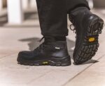 Une personne chaussée de bottes de travail noires dotées de semelles épaisses et profilées se tient debout sur un sol de couleur claire. Les bottes arborent un logo jaune bien visible sur la semelle. Seuls le bas des jambes et les pieds sont visibles.